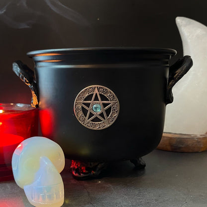 A black 4.5" Pentacle Metal Cauldron adorned with a pentagram emblem is filled with bundled sage. Surrounding the cauldron are an opalescent skull, a large white crystal, orange flowers, sage leaves, and small pyramid-shaped crystals, all set against a dark background.