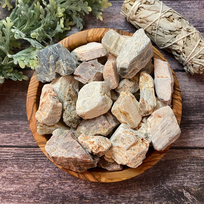 A wooden bowl filled with various rough crystal stones, including Raw Multi Moonstone Tumbled Stones for emotional stability and inner growth, sits on a wooden surface. Next to the bowl, there is a bundle of white sage. Green leafy plants are partially visible in the background.