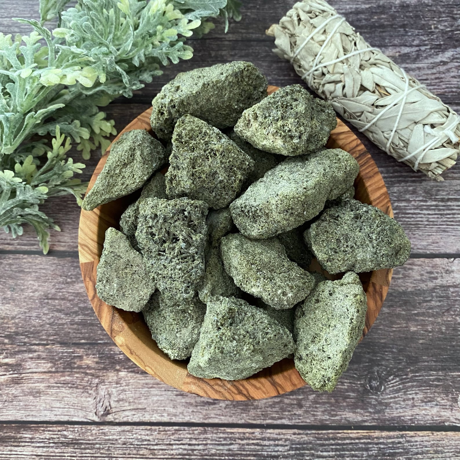 A wooden bowl filled with greenish Raw Epidote Stones on a rustic wooden surface. Beside the bowl, there is a bundle of white sage and some sprigs of greenery, symbolizing spiritual growth.