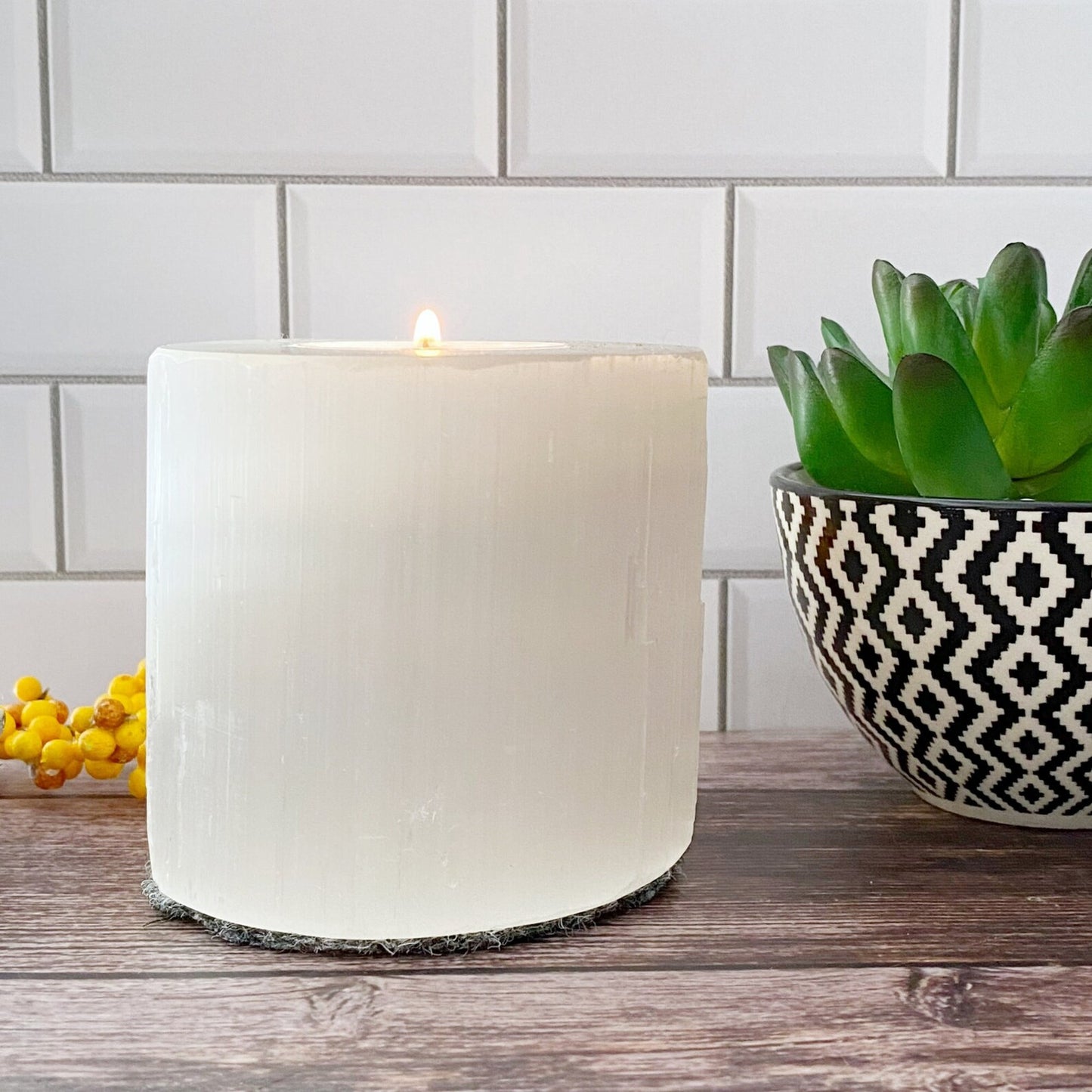 A lit white cylindrical candle stands on a wooden table alongside a 3.25" Selenite Cylinder T-Light Holder, accompanied by a bowl with a black and white geometric pattern holding a green succulent. A cluster of small yellow berries is partially visible to the left, against a white tiled background.