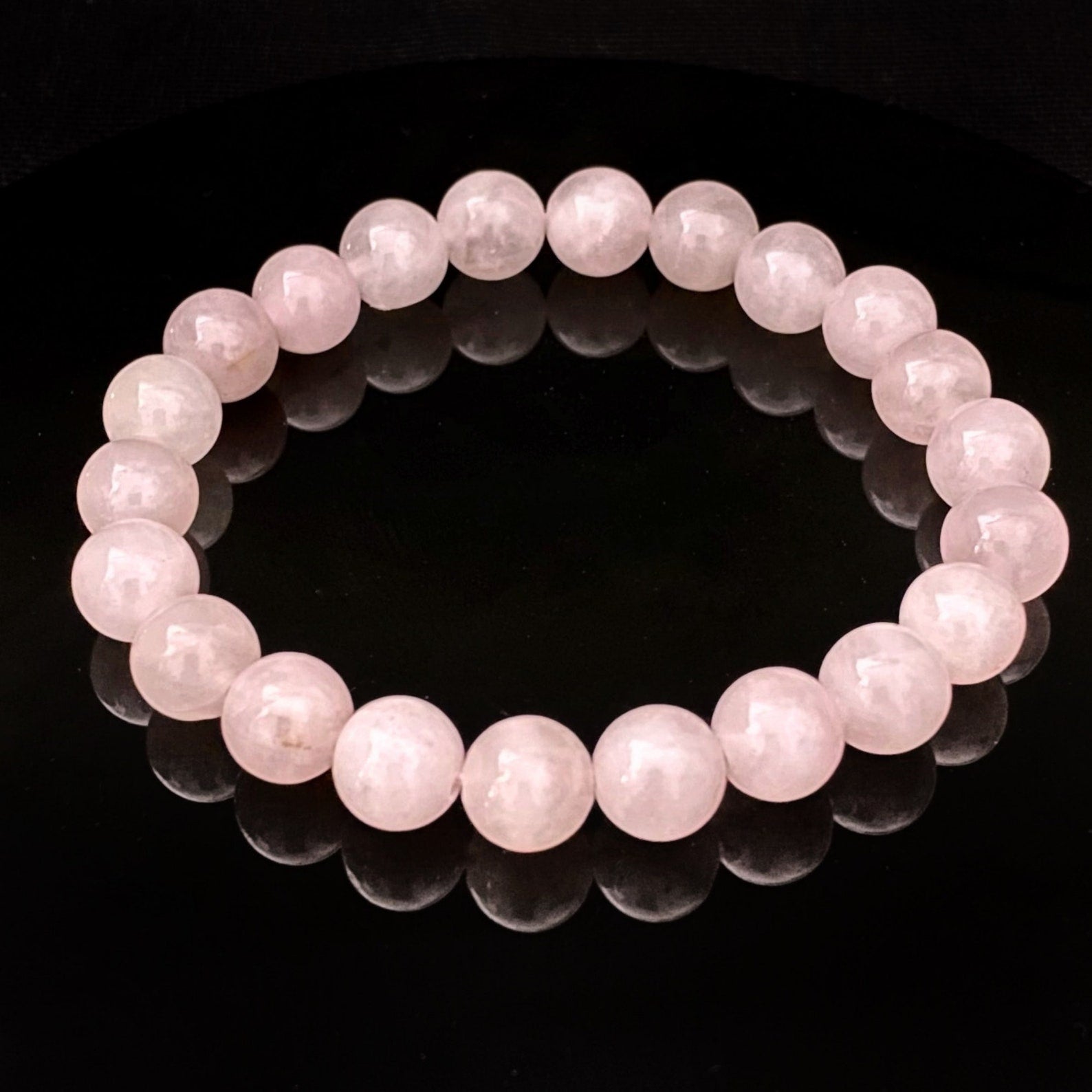 A delicate, light pink 8mm Rose Quartz Beaded Bracelet is showcased against a black background, with its reflection shimmering on the shiny surface below. The bracelet consists of smooth, round beads strung together in a continuous loop, perfect for balancing the Heart Chakra.