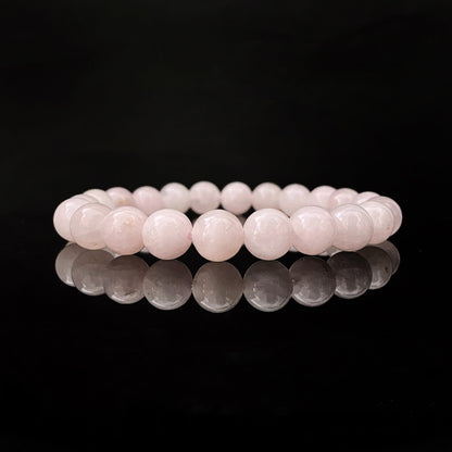 A delicate, light pink 8mm Rose Quartz Beaded Bracelet is showcased against a black background, with its reflection shimmering on the shiny surface below. The bracelet consists of smooth, round beads strung together in a continuous loop, perfect for balancing the Heart Chakra.