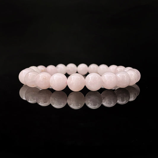 A delicate, light pink 8mm Rose Quartz Beaded Bracelet is showcased against a black background, with its reflection shimmering on the shiny surface below. The bracelet consists of smooth, round beads strung together in a continuous loop, perfect for balancing the Heart Chakra.