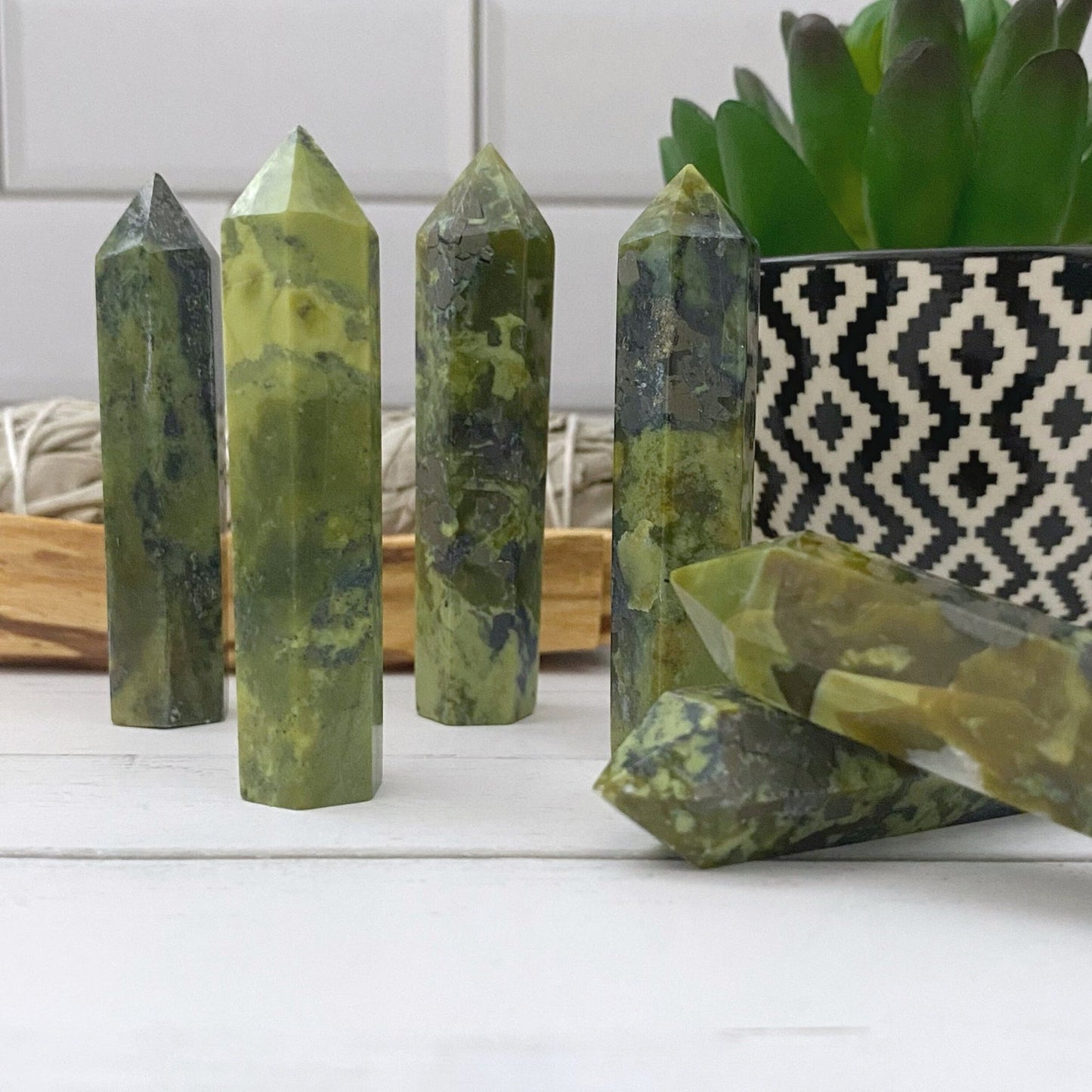 Several polished 2-3" Serpentine Points are arranged upright on a white wooden surface, with one lying down in the foreground. In the background, there's a black and white patterned pot holding a succulent plant, adding an artistic touch that complements these spiritual energy generators.