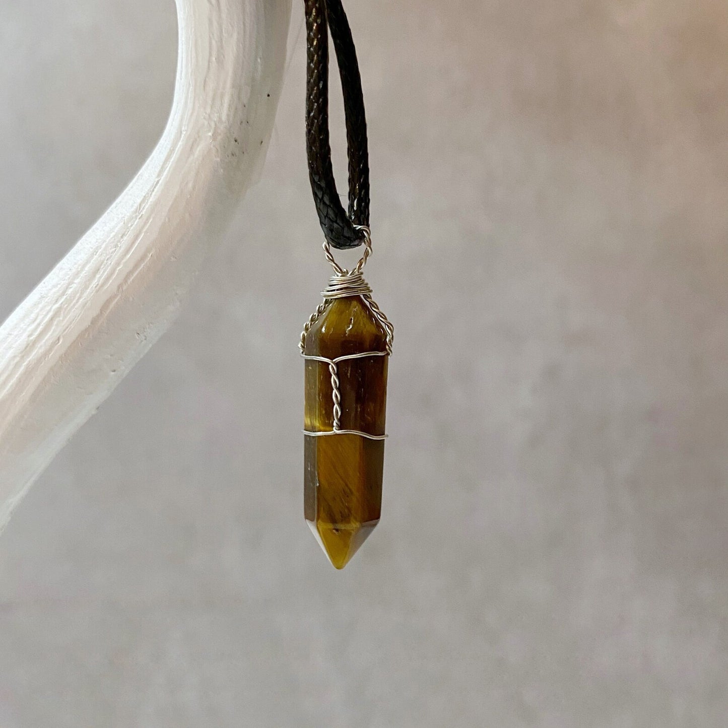 A 1.5" Double Terminated Tiger Eye Point Pendant with a silver wire wrap hangs from a black cotton waxed cord necklace against a neutral background, showcasing its natural brown and yellow stripes.