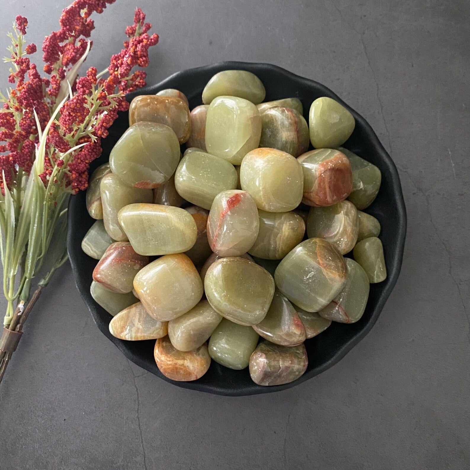 A black bowl filled with polished, greenish-brown stones and Green Aragonite Tumbled Stones is placed on a dark gray surface. To the left of the bowl is a small bunch of red berry-like plant stems with green leaves. These grounding stones have a smooth, glossy finish, perfect for stress relief.