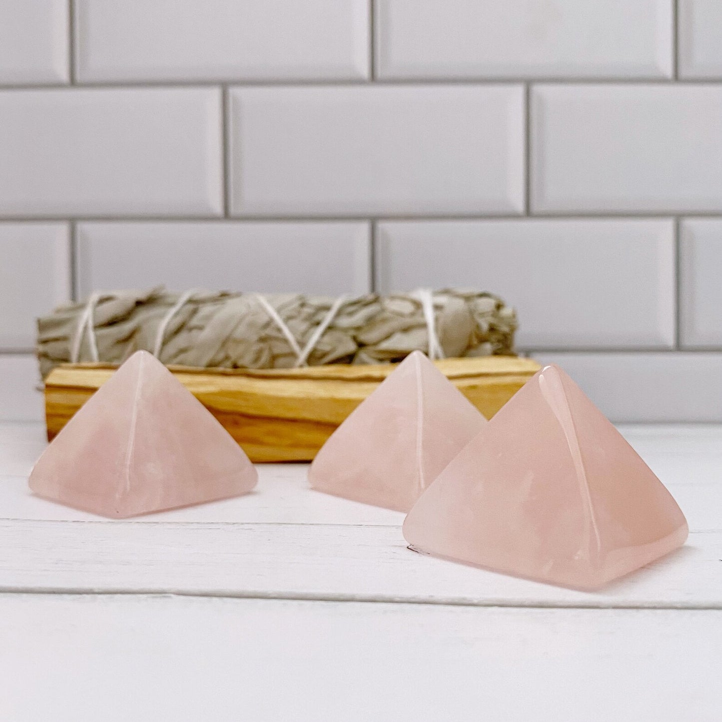 Three pink 1" Mini Rose Quartz Pyramids, known as the Stone of Unconditional Love, are displayed on a white wooden surface in front of a bundled sage smudge stick wrapped in string. The background features white subway tile, adding a clean and serene atmosphere to the composition.