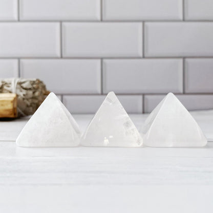 Three 1" Mini Clear Quartz Pyramids are lined up on a white surface in front of a white subway tile wall, radiating serenity energy. In the background, a bundle of dried sage rests on a wooden holder. The setting appears minimalistic and serene, perfect for creating a meditation area.