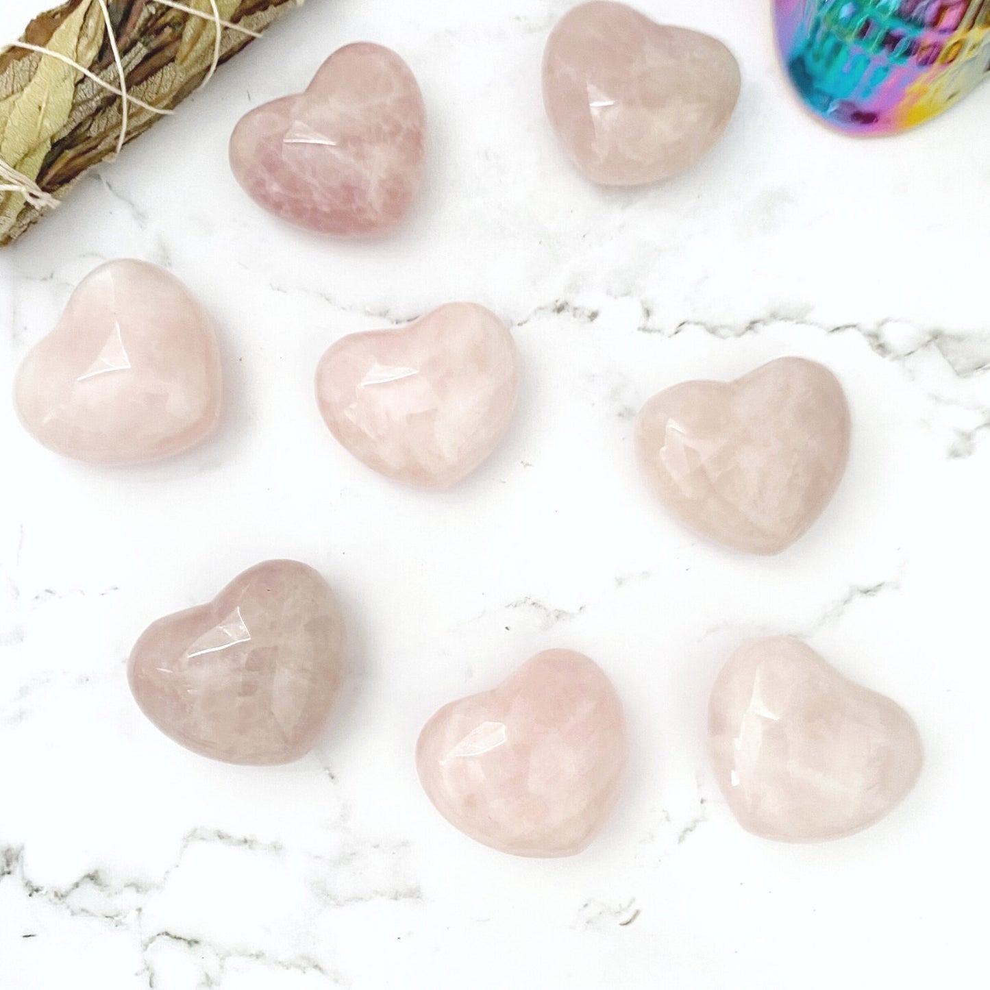 Eight 2" Puffy Rose Quartz Palm Hearts, known as the Stone of Unconditional Love, are arranged on a white marble surface. To the left is a bundle of sage, and in the top right corner, there's a small, colorful, iridescent container. The image radiates emotional healing with its soft, calming aesthetic.