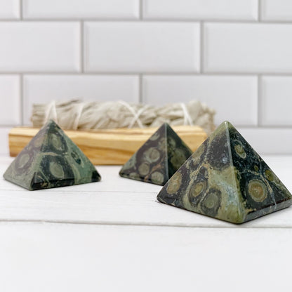 Three 1" Mini Kambaba Jasper Pyramids with calming green and black patterns are placed on a white surface. In the background, there is a bundle of sage tied with string, set against a white tiled wall. The scene is well-lit and has a clean, minimalist aesthetic.