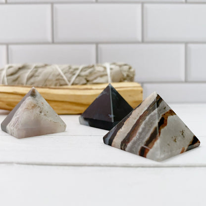 Three 1" Mini Sardonyx Pyramids with varying colors and patterns sit on a white surface in the foreground. In the background, there is a bundle of sage resting in a small wooden tray. The backdrop consists of white subway tiles, creating an elegant setting for this unique crystal collection.