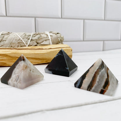 Three 1" Mini Sardonyx Pyramids with varying colors and patterns sit on a white surface in the foreground. In the background, there is a bundle of sage resting in a small wooden tray. The backdrop consists of white subway tiles, creating an elegant setting for this unique crystal collection.