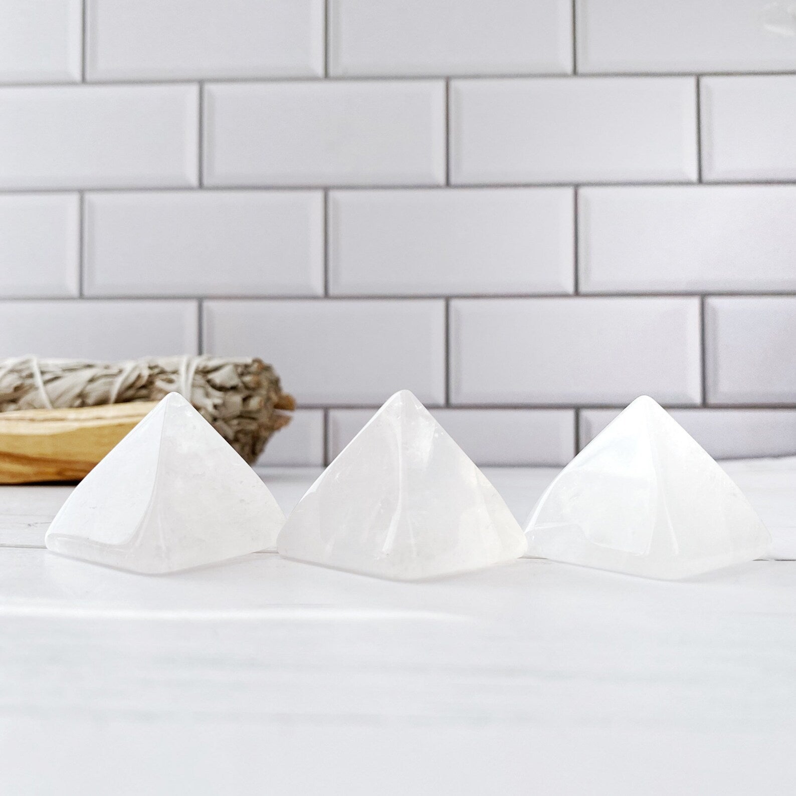 Three 1" Mini Clear Quartz Pyramids are lined up on a white surface in front of a white subway tile wall, radiating serenity energy. In the background, a bundle of dried sage rests on a wooden holder. The setting appears minimalistic and serene, perfect for creating a meditation area.