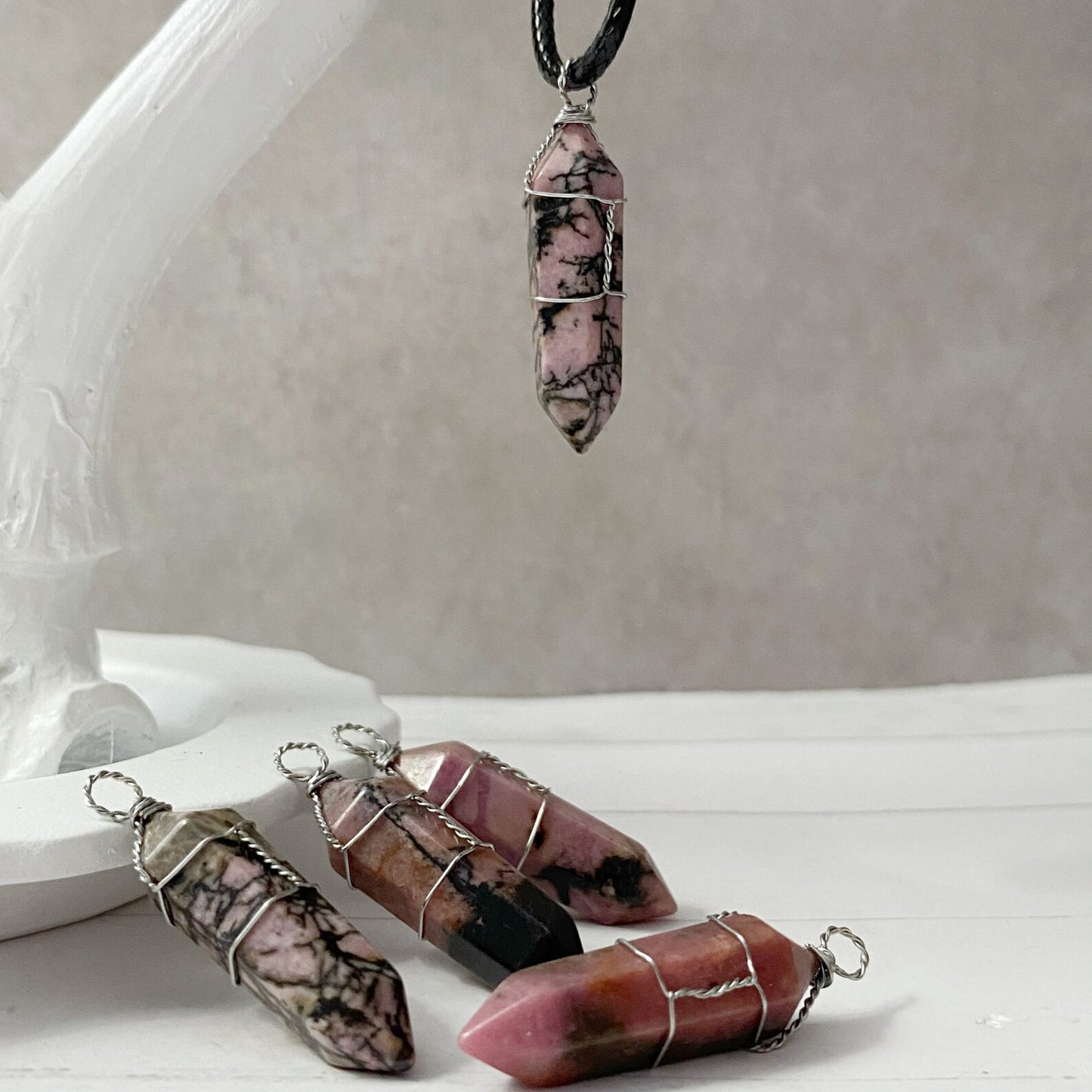 A 1.5" Double Terminated Rhodonite Point Pendant, crafted from pink and black marbled stone, is attached to a thin black cotton waxed cord necklace. The pendant, wire-wrapped with silver-colored metallic accents, is displayed on a white textured background.