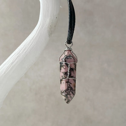 A 1.5" Double Terminated Rhodonite Point Pendant, crafted from pink and black marbled stone, is attached to a thin black cotton waxed cord necklace. The pendant, wire-wrapped with silver-colored metallic accents, is displayed on a white textured background.