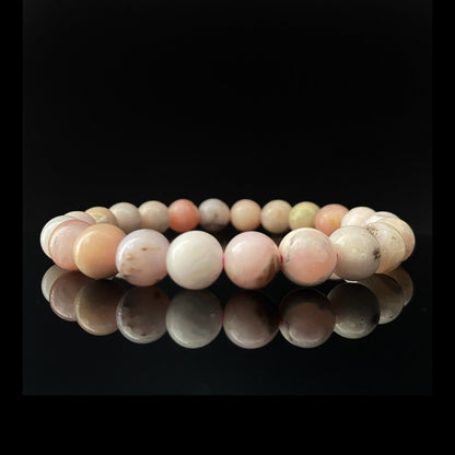 A close-up shot of an 8mm Pink Opal Beaded Bracelet made of polished, multicolored stones, with hues of pink, white, and hints of green. The bracelet is placed against a black background, with its reflection visible on a glossy surface below, symbolizing heart chakra balance and emotional healing.