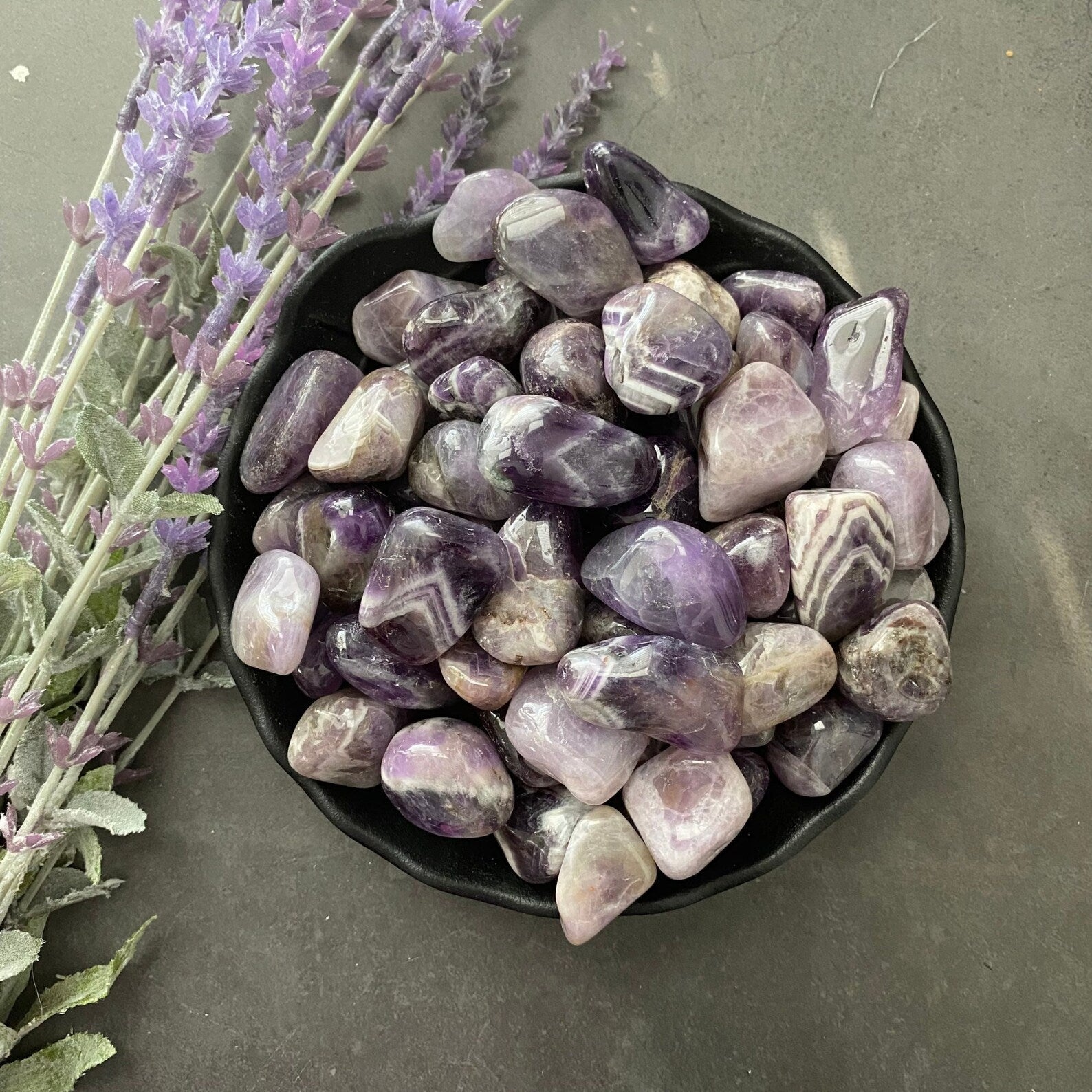 A black bowl filled with polished Chevron Amethyst Tumbled Stones, varying in shades of purple and white, is placed on a dark surface. Purple lavender sprigs are arranged next to the bowl, providing a soothing and natural contrast while enhancing intuition with their stress-relieving qualities.