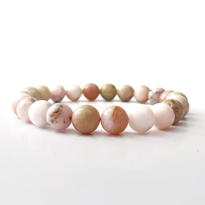 A close-up shot of an 8mm Pink Opal Beaded Bracelet made of polished, multicolored stones, with hues of pink, white, and hints of green. The bracelet is placed against a black background, with its reflection visible on a glossy surface below, symbolizing heart chakra balance and emotional healing.