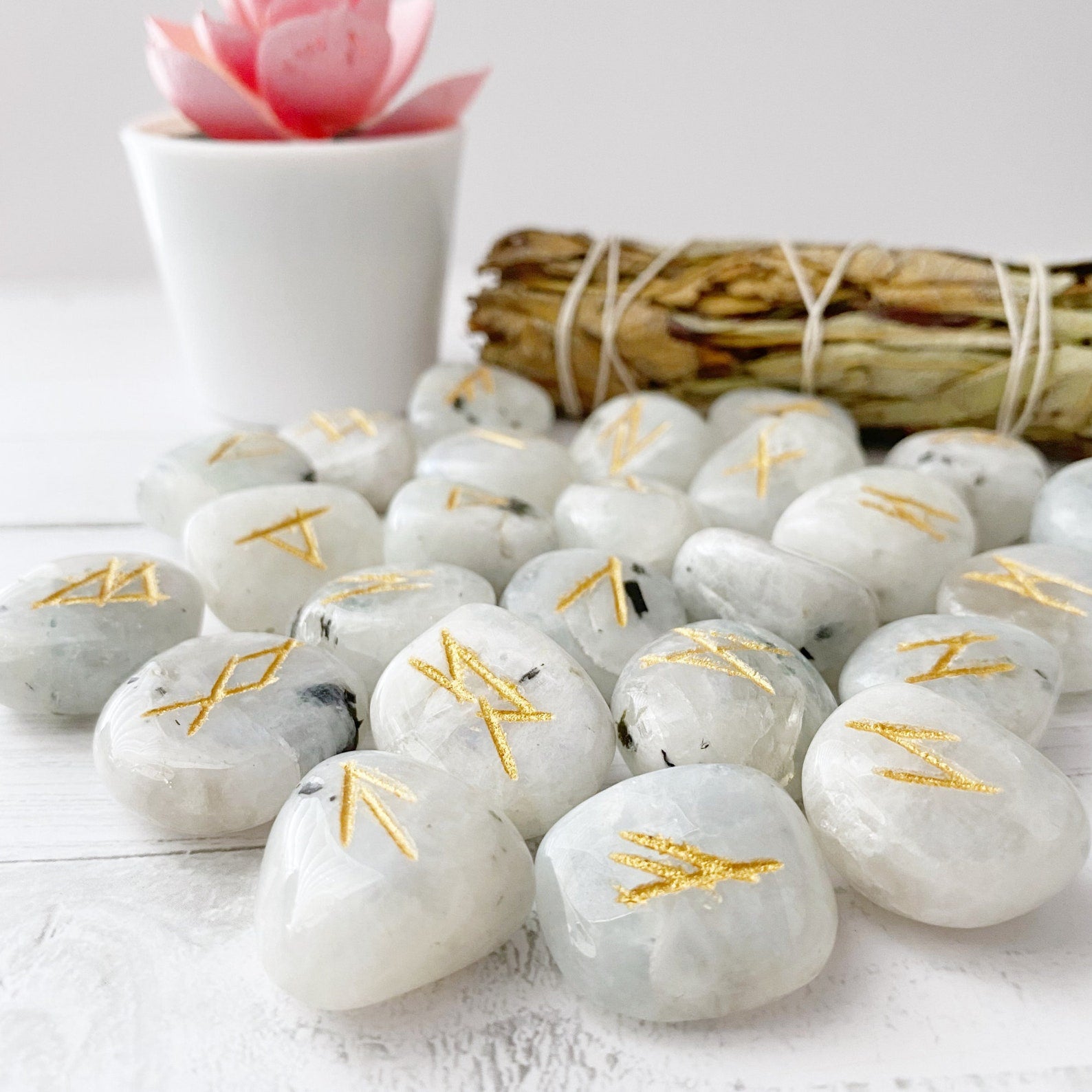 Smooth, white Rainbow Moonstone Elder Futhark Runes with gold engravings are scattered on a white wooden surface. In the background, a small potted plant with a pink flower and a bundle of dried sage leaves tied together are visible.