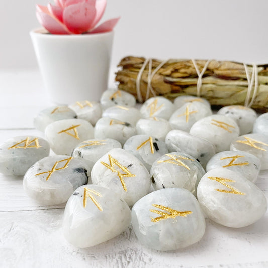 Smooth, white Rainbow Moonstone Elder Futhark Runes with gold engravings are scattered on a white wooden surface. In the background, a small potted plant with a pink flower and a bundle of dried sage leaves tied together are visible.