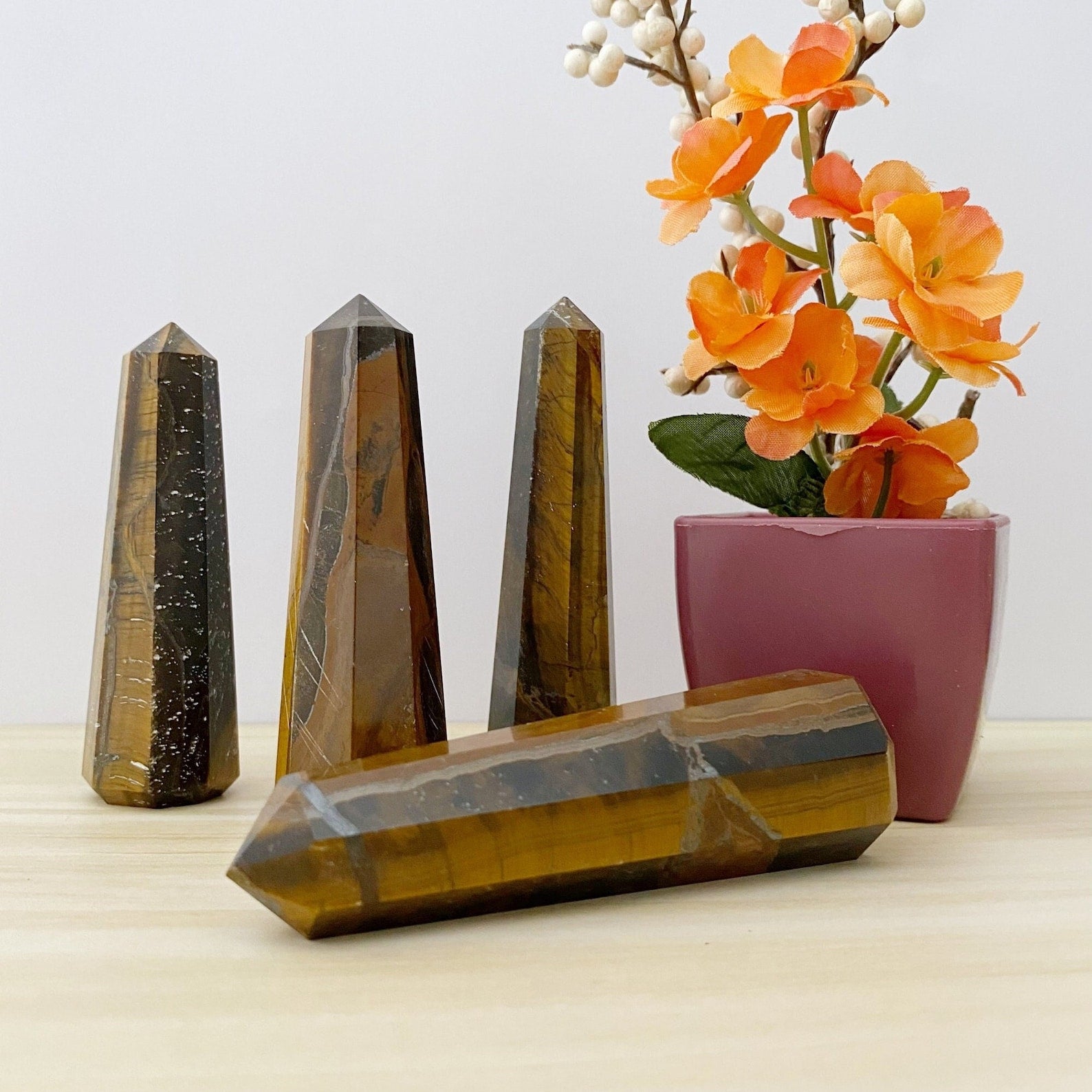 A group of four polished 3-4" Tiger Eye Points in various sizes stand on a wooden surface next to a small pink pot containing vibrant orange and white artificial flowers. One point is lying horizontally in front of the others, resembling an elegant energy generator.