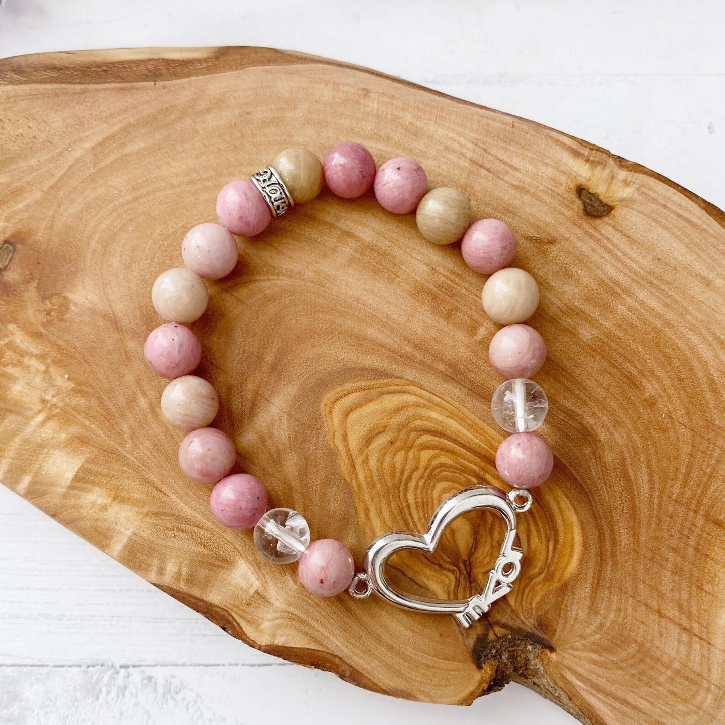 An 8mm Rhodonite & Quartz Beaded Bracelet with Heart, featuring pink and beige stones and a silver heart-shaped clasp, rests on a wooden surface. In the background, there is a blurry sprig of purple lavender, enhancing its aura.