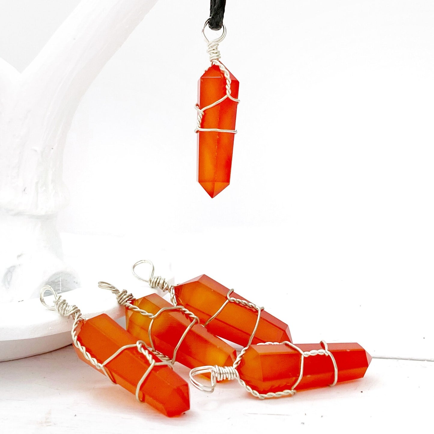 A vibrant orange 1.5" Double Terminated Carnelian Point Pendant, intricately wrapped in silver wire, hangs on a black cord against a white background. The faceted, pointed crystal is held securely by the wire, displaying a polished and glossy finish and resonating with sacral chakra energy.