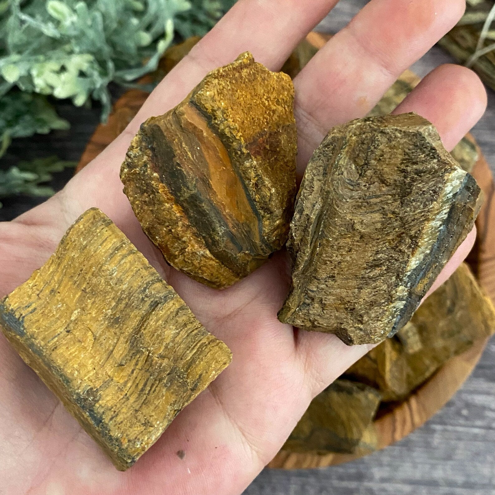 A wooden bowl filled with raw unpolished Raw Tiger Eye Stones sits on a wooden surface. To the left, green leafy foliage is partially visible, and a bundled sage stick rests above the bowl to the right. The background features a rustic wooden texture, perfect for inspiring your next crafting pendants project.