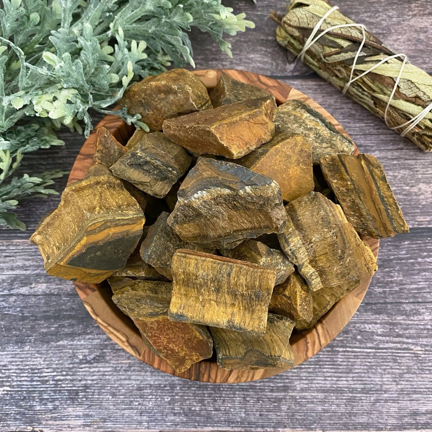 A wooden bowl filled with raw unpolished Raw Tiger Eye Stones sits on a wooden surface. To the left, green leafy foliage is partially visible, and a bundled sage stick rests above the bowl to the right. The background features a rustic wooden texture, perfect for inspiring your next crafting pendants project.