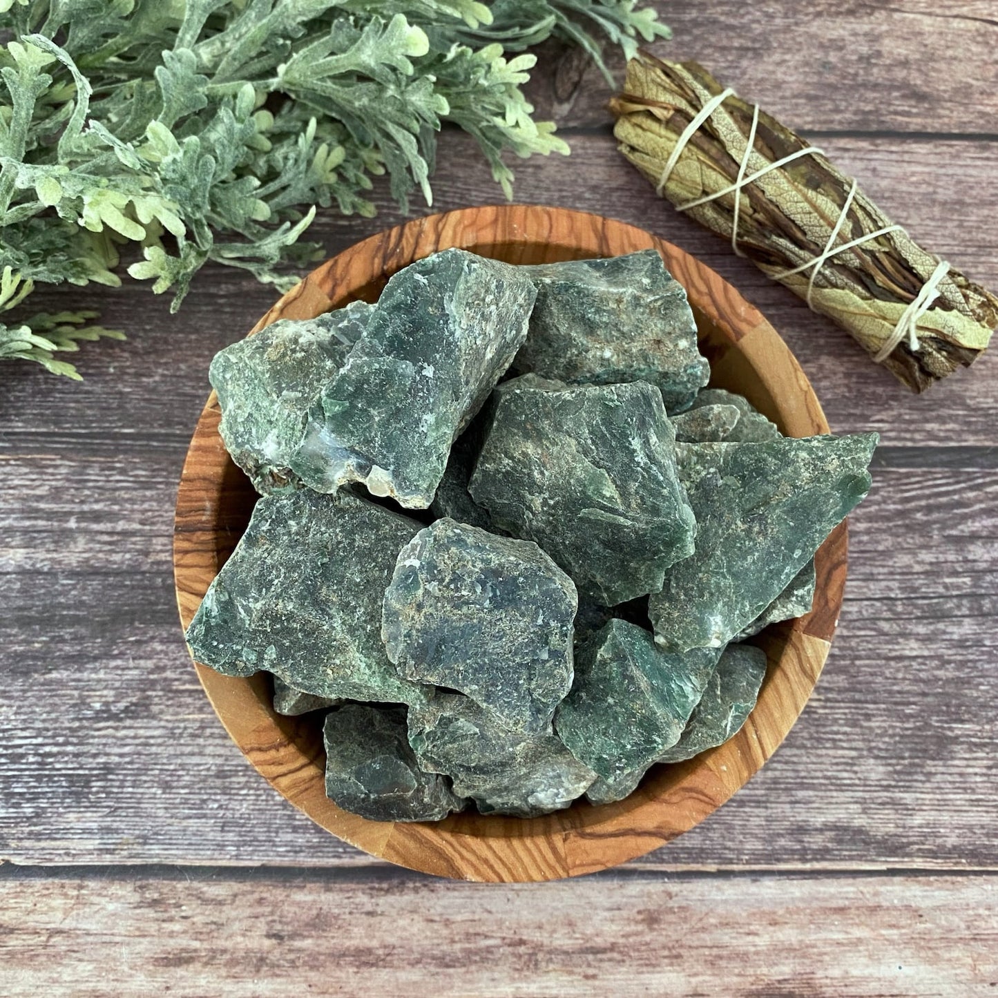 A wooden bowl filled with large, rough Raw Green Moss Agate Stones is placed on a wooden surface. Beside it are sprigs of greenery and a small bundle of dried herbs tied together with string, perfect for energy work.