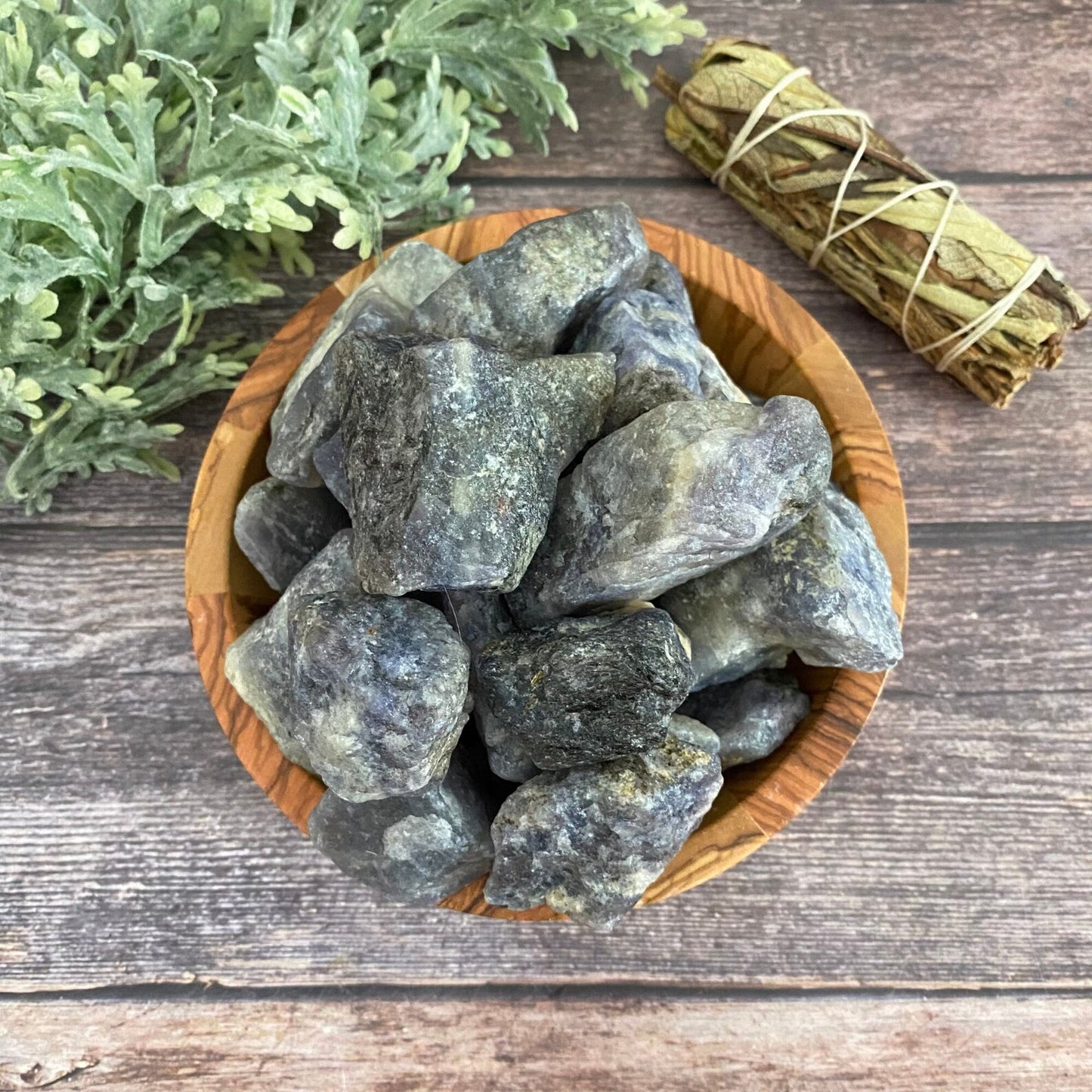 A wooden bowl filled with large, rough-cut Raw Iolite Stones sits on a rustic wooden surface. Beside the bowl, there is a bundle of dried sage wrapped with string. In the background, there is a leafy green plant.