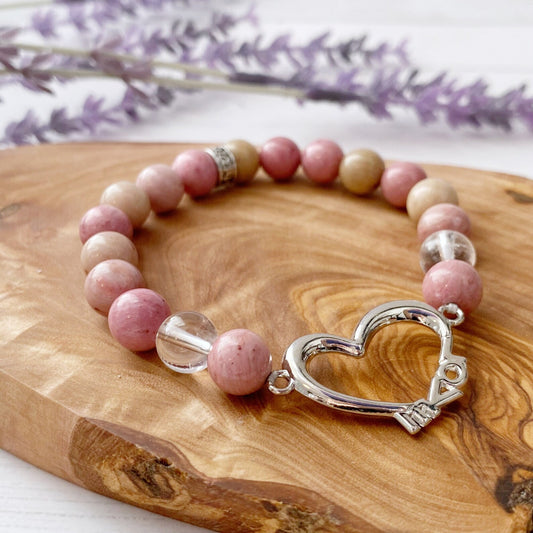 An 8mm Rhodonite & Quartz Beaded Bracelet with Heart, featuring pink and beige stones and a silver heart-shaped clasp, rests on a wooden surface. In the background, there is a blurry sprig of purple lavender, enhancing its aura.