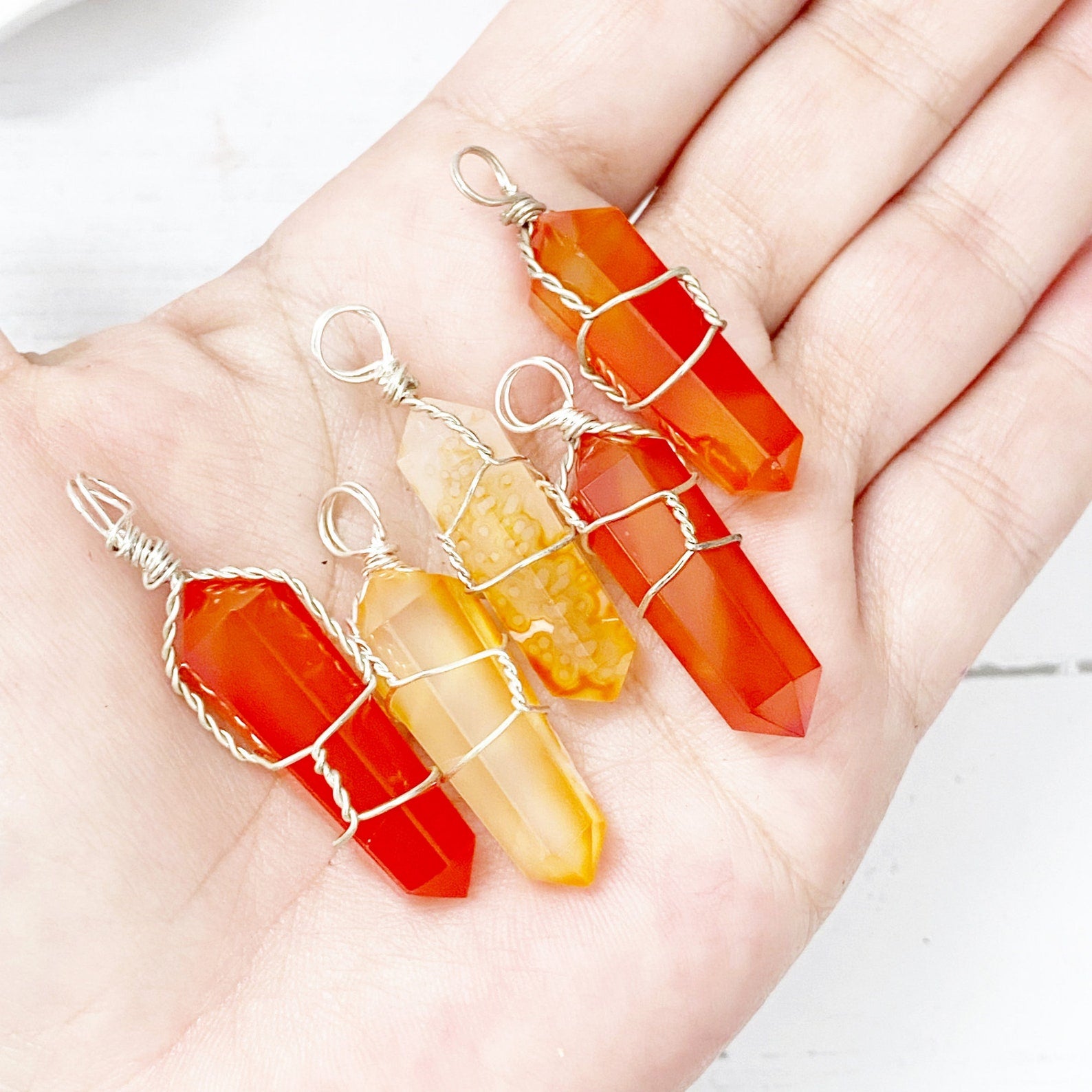 A vibrant orange 1.5" Double Terminated Carnelian Point Pendant, intricately wrapped in silver wire, hangs on a black cord against a white background. The faceted, pointed crystal is held securely by the wire, displaying a polished and glossy finish and resonating with sacral chakra energy.