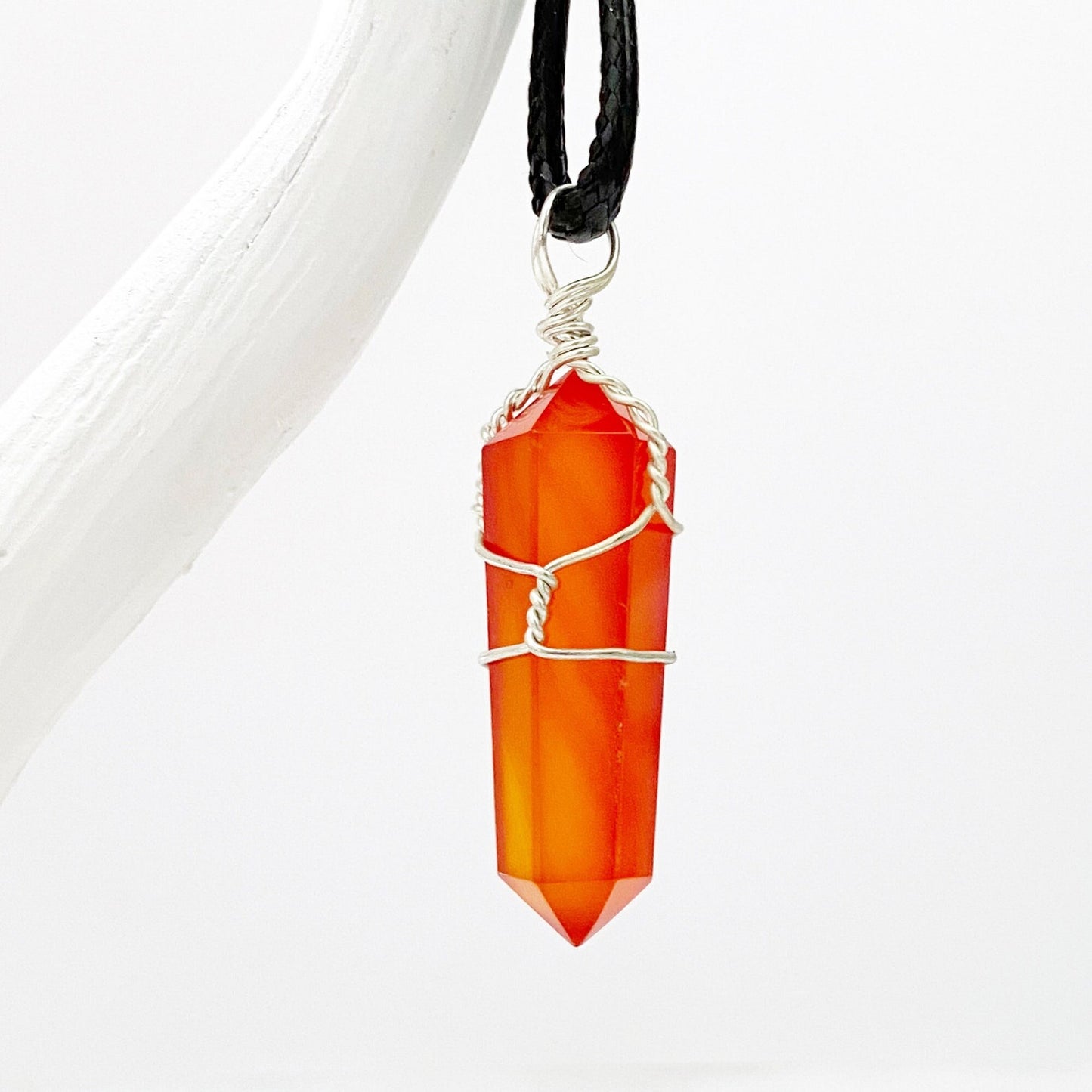 A vibrant orange 1.5" Double Terminated Carnelian Point Pendant, intricately wrapped in silver wire, hangs on a black cord against a white background. The faceted, pointed crystal is held securely by the wire, displaying a polished and glossy finish and resonating with sacral chakra energy.