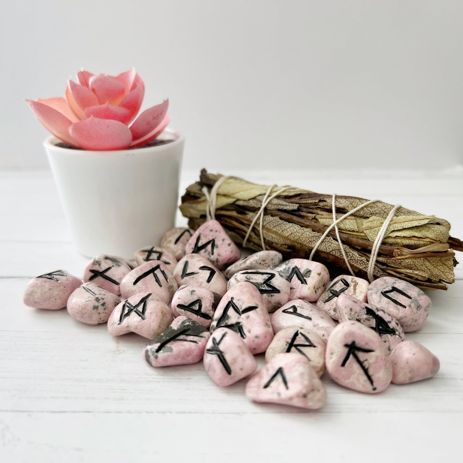 A white table holds a small pink succulent in a white pot, a bound bundle of dried sage, and a collection of Rhodonite Elder Futhark Runes. The setting is clean with a minimalist aesthetic.