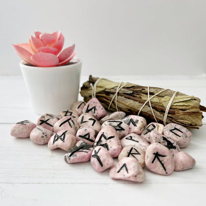 A white table holds a small pink succulent in a white pot, a bound bundle of dried sage, and a collection of Rhodonite Elder Futhark Runes. The setting is clean with a minimalist aesthetic.