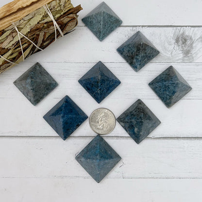 A collection of smooth, 1" Mini Dumortierite Pyramids, known for enhancing psychic abilities and Third Eye Chakra activation, are displayed on a white surface with a white tiled wall in the background. A basket of coiled rope is partially visible in the background. The focus is on a central pyramid in the foreground.