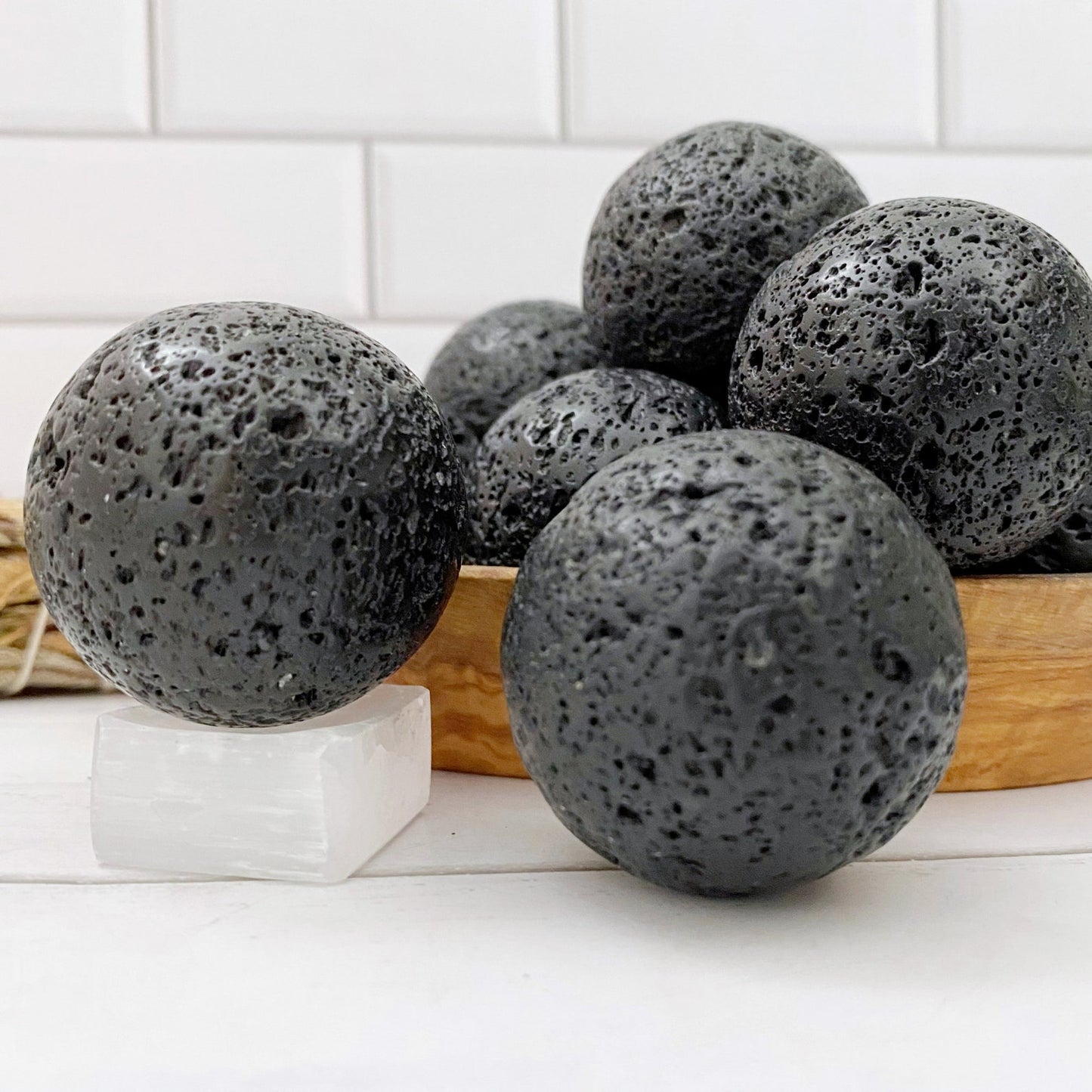 A close-up of six black, porous bath bombs arranged in a wooden bowl on a white tiled background. One bath bomb, resembling a 40mm Lava Rock Crystal Ball, is placed separately on a small clear block in the foreground, highlighting its textured surface and grounding earth energy.