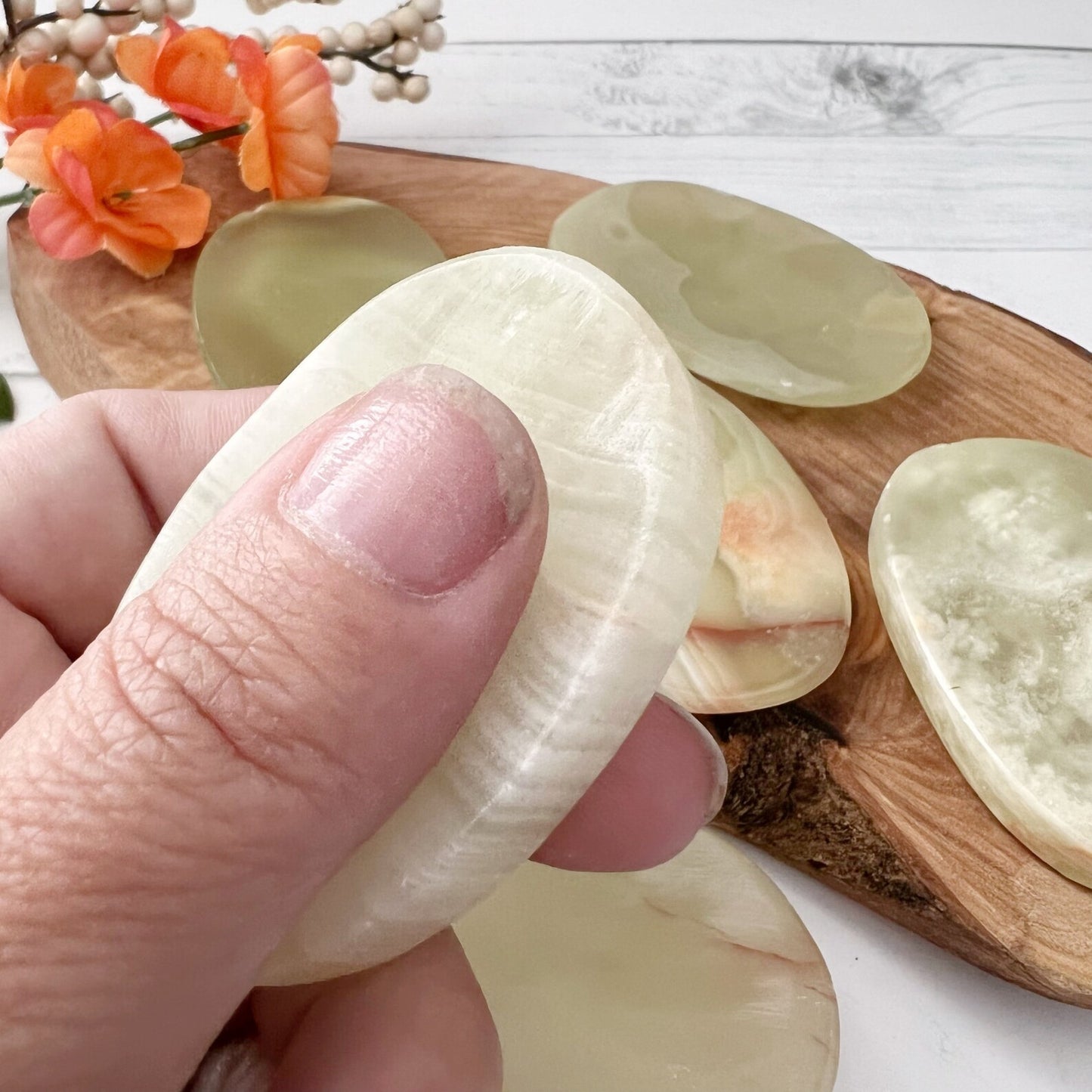 A wooden tray holds four polished, oval-shaped Green Onyx Worry Stones of varying translucency and patterns, known for their heart chakra properties. Orange flowers in the top left corner add a touch of color to the composition. The scene is set on a white surface.