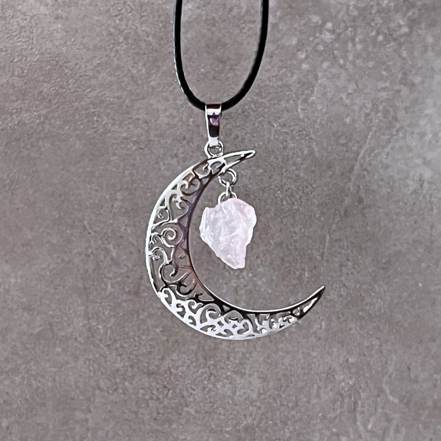 A Silver Crescent Moon & Rose Quartz Pendant features intricate filigree detailing and a rough-cut Rose Quartz crystal hanging from its top point. The pendant is attached to a black cotton-waxed cord necklace, displayed against a textured gray background.