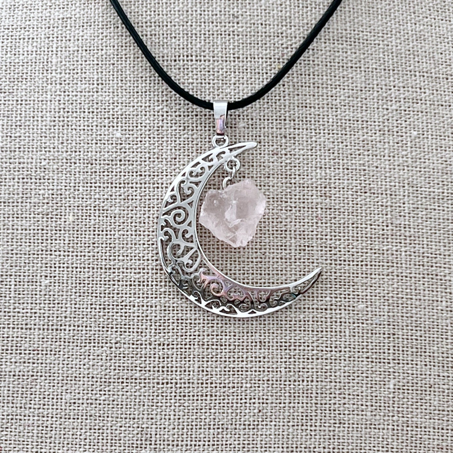 A Silver Crescent Moon & Rose Quartz Pendant features intricate filigree detailing and a rough-cut Rose Quartz crystal hanging from its top point. The pendant is attached to a black cotton-waxed cord necklace, displayed against a textured gray background.