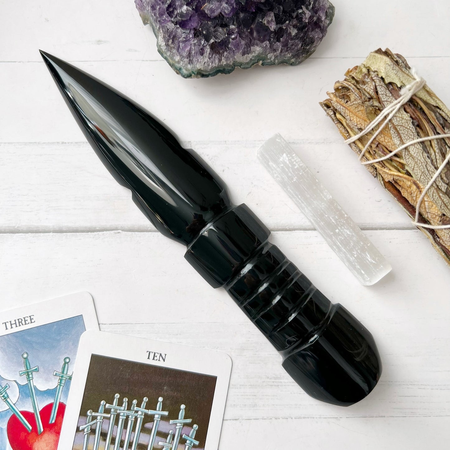 A 7"-8" Black Obsidian Athame is placed on a white wooden surface. Surrounding it are an amethyst cluster, a sage bundle, a selenite stick, and two tarot cards featuring the Three of Swords and Ten of Swords.