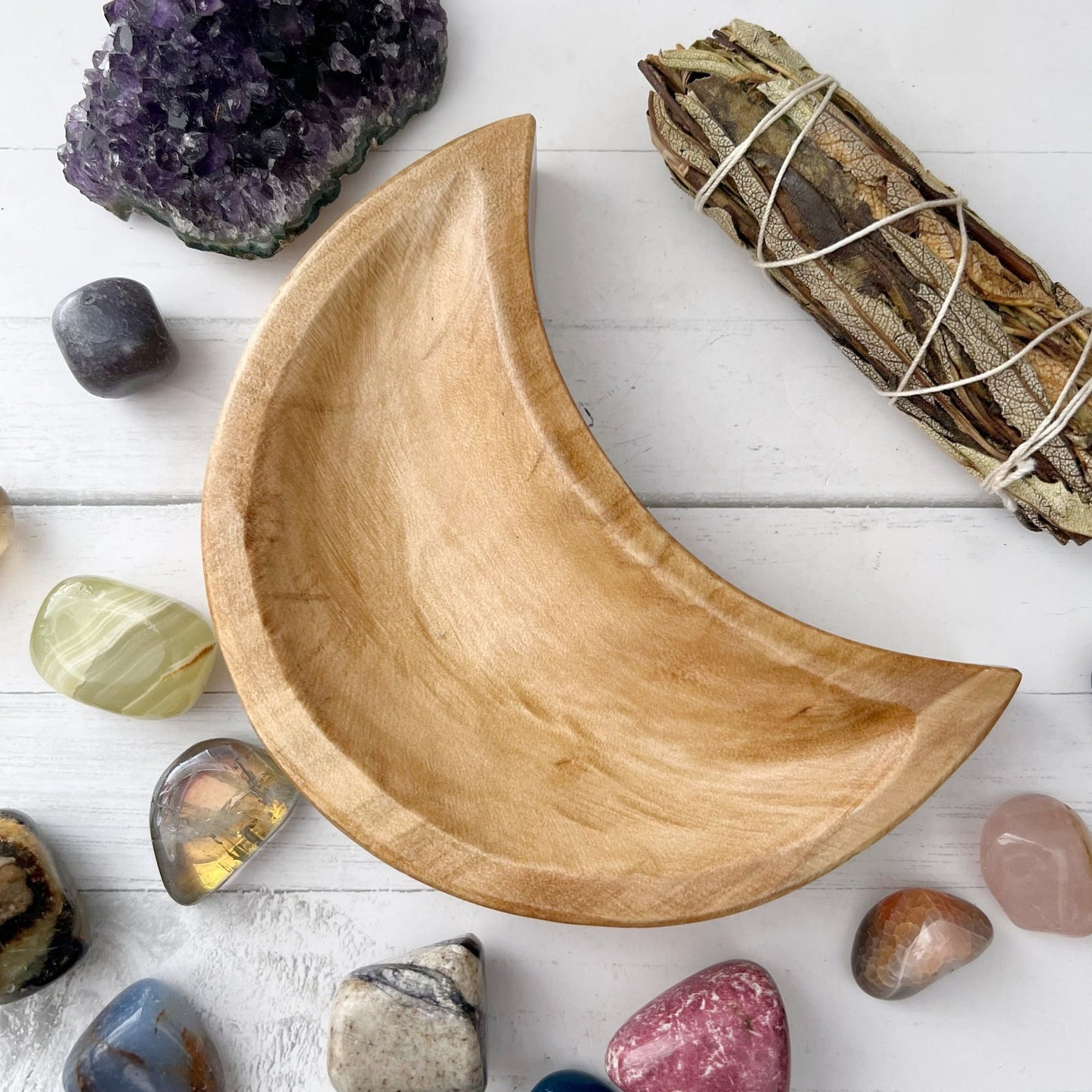 A 5" Crescent Moon Teak Wood Bowl is surrounded by various polished gemstones, a cluster of purple crystals, and a bundle of dried sage on a white surface—perfect for store crystals or metaphysical work.