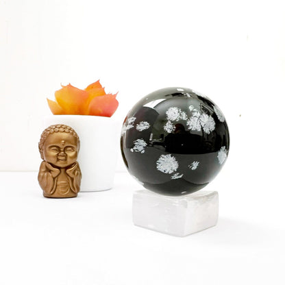 A polished black sphere of 40mm Snowflake Obsidian Crystal Ball with white floral-like patterns is displayed on a square white stand. To its left, a small bronze-colored Buddha statue sits with a serene expression, and behind it, there is a white pot containing a succulent with orange-tipped leaves.