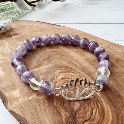 A bracelet made of round 8mm Chevron Amethyst Beaded Bracelet with Lotus Charm stones with Clear Quartz beads and a silver lotus flower charm. The bracelet is resting on a wooden surface with green fabric and white flowers in the background, emanating an aura of Usui Reiki healing.