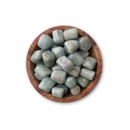 A wooden bowl filled with polished, light blue-green Aquamarine Tumbled Stones, each stone smooth and rounded, resting against one another. The bowl's natural wooden grain contrasts with the smooth texture of the stones, perfect for chakra alignment. The background is white.