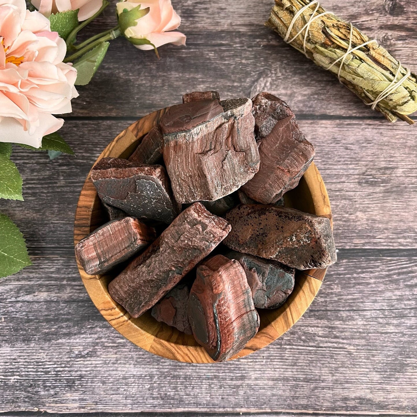 A wooden bowl filled with Raw Red Tiger Eye Stones, perfect for spiritual practices or jewelry making, is placed on a wooden surface. Next to the bowl are some pink flowers and a bundle of dried sage wrapped with string.