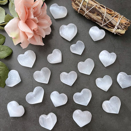 A collection of small, polished, white 1" Mini Selenite Pocket Hearts arranged on a dark surface exudes spiritual balance. A peach-colored flower and a bound bundle of dried herbs rest at the top left of the image, flanked by green leaves, enhancing the sense of mental clarity and harmony.