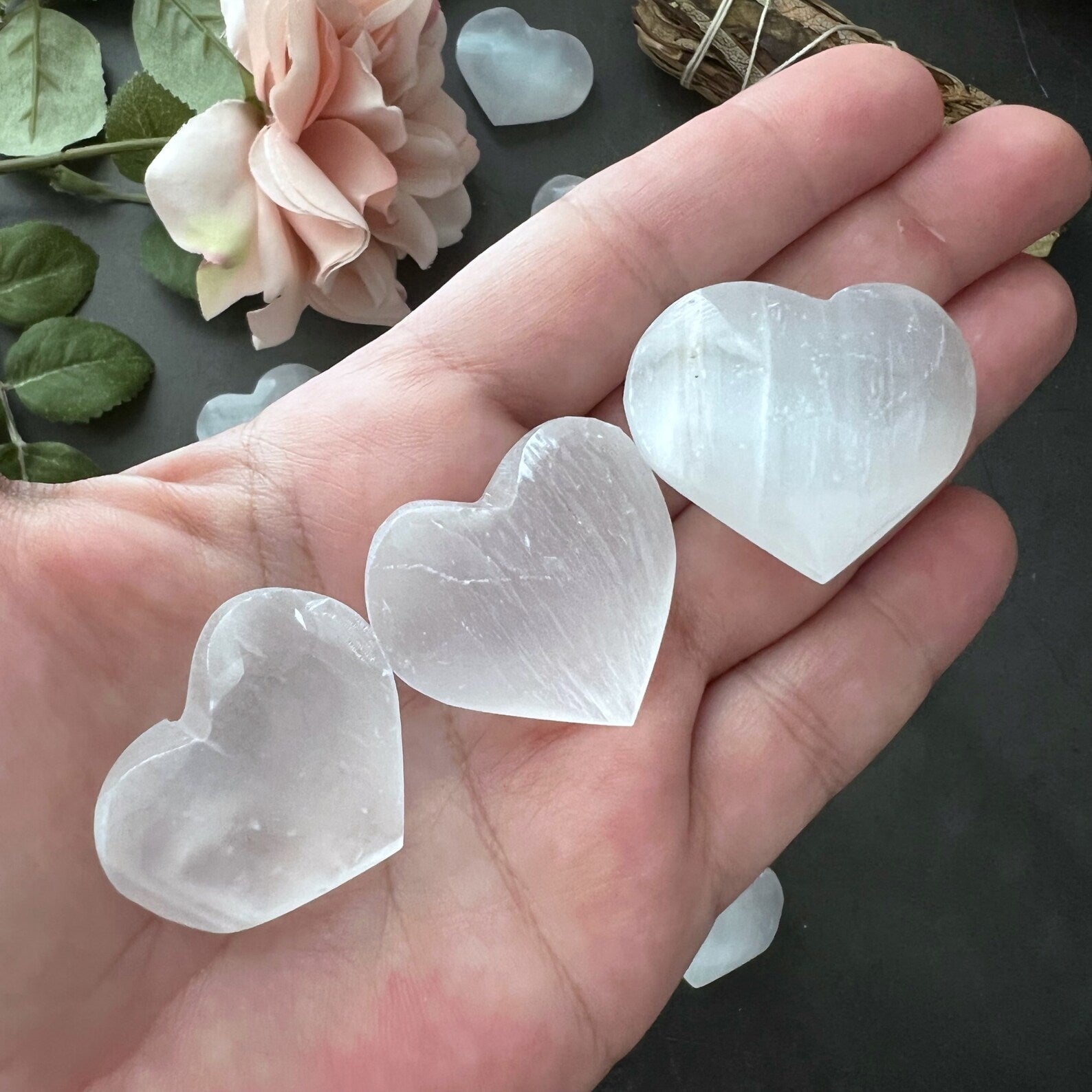 A collection of small, polished, white 1" Mini Selenite Pocket Hearts arranged on a dark surface exudes spiritual balance. A peach-colored flower and a bound bundle of dried herbs rest at the top left of the image, flanked by green leaves, enhancing the sense of mental clarity and harmony.