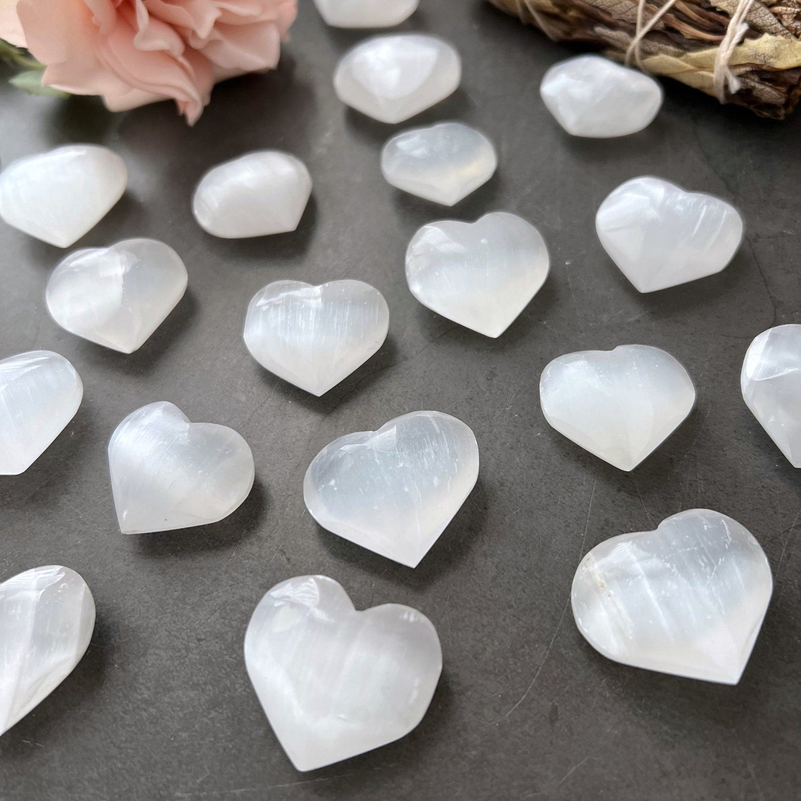 A collection of small, polished, white 1" Mini Selenite Pocket Hearts arranged on a dark surface exudes spiritual balance. A peach-colored flower and a bound bundle of dried herbs rest at the top left of the image, flanked by green leaves, enhancing the sense of mental clarity and harmony.